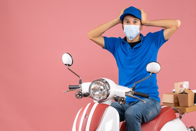 Vista superior del chico de entrega emocional confundido en máscara médica con sombrero sentado en scooter sobre fondo de melocotón pastel