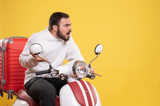 Vista superior del chico joven sentado en la motocicleta con la maleta y mirando algo con expresión facial sorprendida sobre fondo amarillo aislado