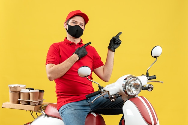 Vista superior del chico de mensajería con blusa roja y guantes de sombrero en máscara médica entregando orden sentado en scooter apuntando hacia arriba