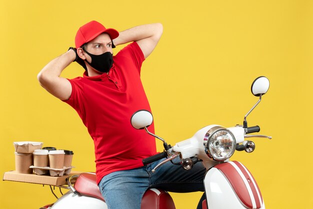 Vista superior del chico de mensajería con blusa roja y guantes de sombrero en máscara médica entregando orden sentado en scooter sintiéndose confundido