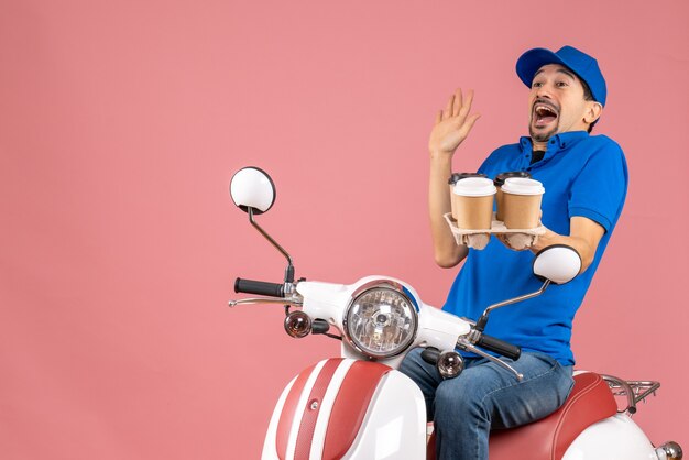 Vista superior del chico de mensajero sorprendido en máscara médica con sombrero sentado en scooter con orden en melocotón pastel