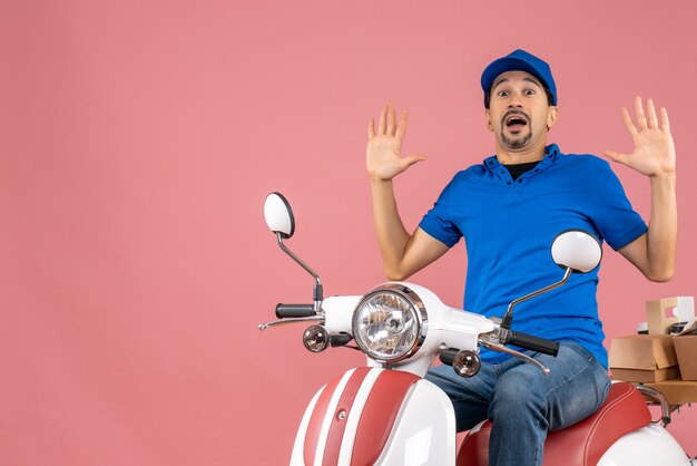 Vista superior del chico mensajero sorprendido con sombrero sentado en scooter entregando pedidos sobre fondo melocotón pastel