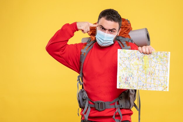Vista superior de un chico viajero pensativo emocional con máscara médica con mochila con mapa sobre fondo amarillo