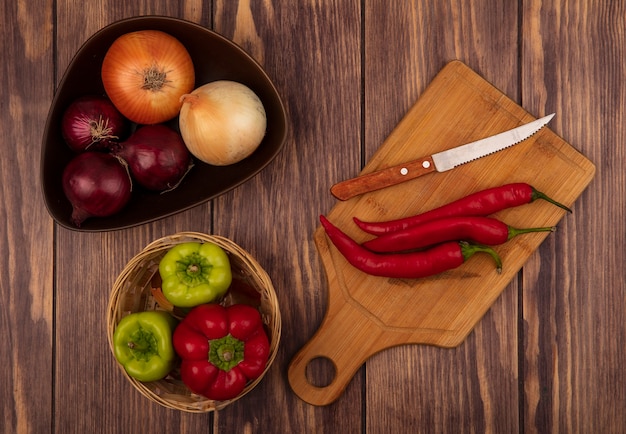 Vista superior de chiles frescos en una tabla de cocina de madera con cuchillo con cebollas en un recipiente con pimientos en un cubo en una pared de madera