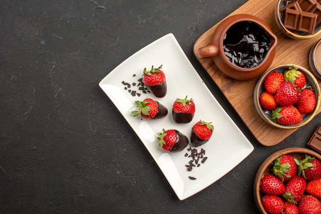Vista superior de chocolate a bordo de barras de chocolate junto a la tabla de cortar con crema de chocolate y fresas y fresas cubiertas de chocolate en un plato