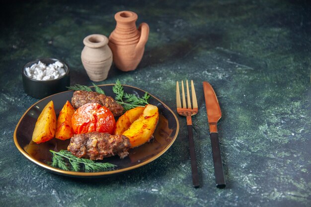 Vista superior de chuletas de carne al horno con patatas y tomate servido con cubiertos verde sal sobre fondo de color oscuro