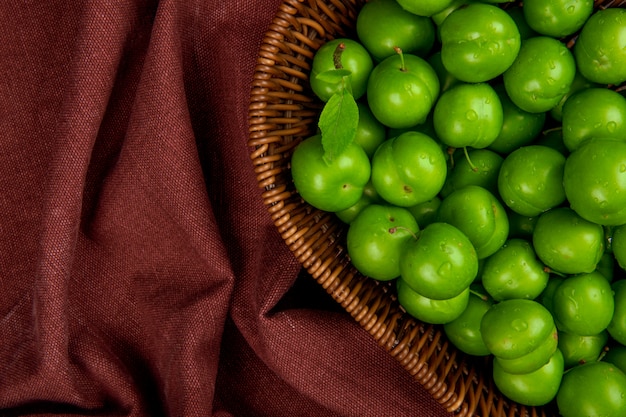 Foto gratuita vista superior de ciruelas agrias verdes en una cesta de mimbre sobre una mesa de tela rojo oscuro con espacio de copia