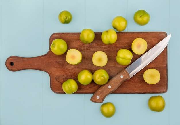 Vista superior de ciruelas cerezas verdes frescas en una tabla de cocina de madera con cuchillo sobre un fondo azul.