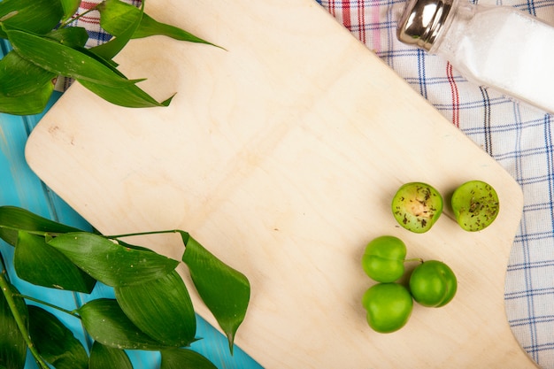 Vista superior de ciruelas verdes agrias y un salero en una tabla de cortar de madera con hojas de ruscus en una mesa de tela a cuadros