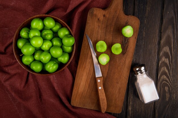 Vista superior de ciruelas verdes agrias en un tazón y ciruelas en rodajas sobre una tabla de cortar de madera con cuchillo y un salero en la mesa rústica oscura