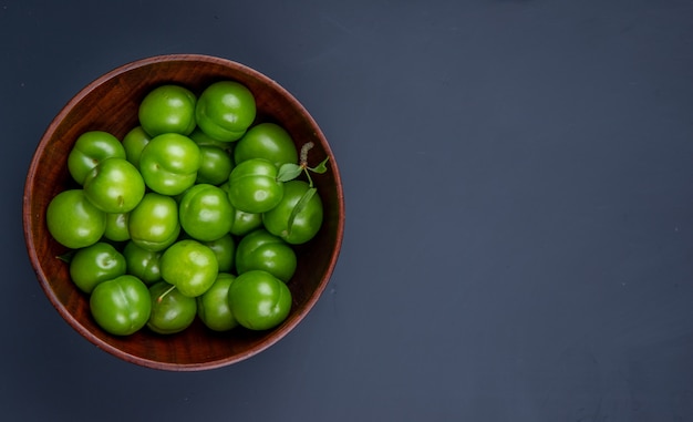 Vista superior de ciruelas verdes agrias en un tazón de madera sobre una mesa negra con espacio de copia