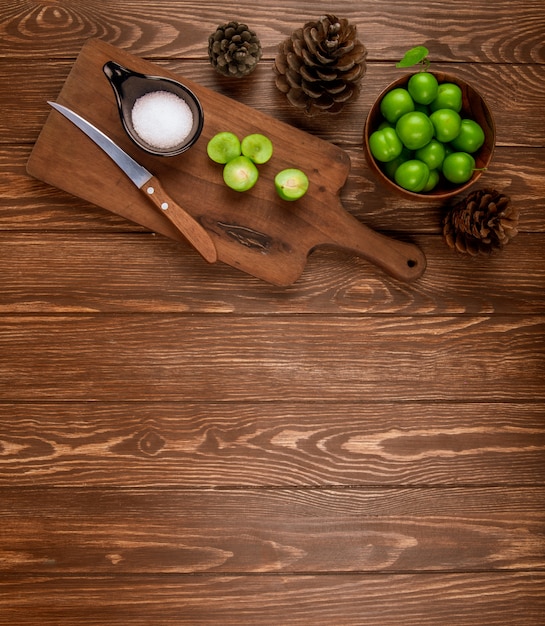 Vista superior de ciruelas verdes en rodajas con sal y cuchillo de cocina en una tabla de cortar de madera, conos y ciruelas en un recipiente en la mesa de madera rústica con espacio de copia