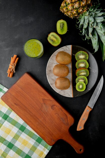 Vista superior de cítricos como kiwi y piña con tabla para cortar canela y cuchillo sobre superficie negra