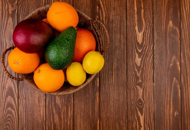 Vista superior de cítricos como mango naranja aguacate limón en canasta en el lado izquierdo y fondo de madera con espacio de copia