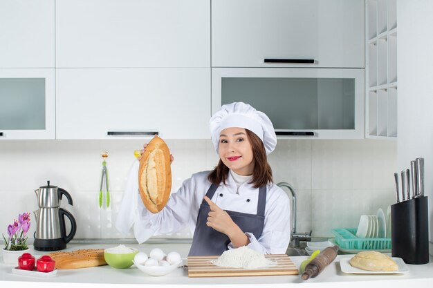 Vista superior de la cocinera en uniforme de pie detrás de la mesa con alimentos de tabla de cortar sosteniendo y señalando el pan en la cocina blanca