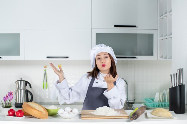Vista superior de la cocinera en uniforme de pie detrás de la mesa con verduras de pan de tabla de cortar enviando gesto de beso en la cocina blanca