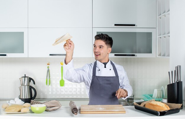 Vista superior del cocinero joven sonriente en uniforme de pie detrás de la mesa en la cocina blanca