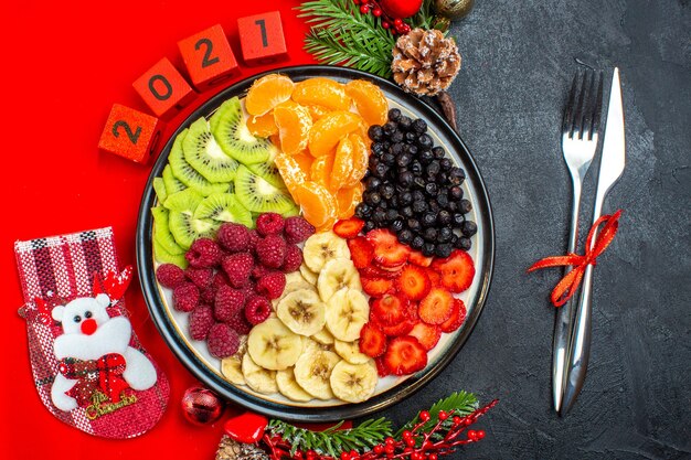 Vista superior de la colección de frutas frescas en accesorios de decoración de plato de cena ramas de abeto y números calcetín de Navidad en una servilleta roja y cubiertos sobre un fondo negro