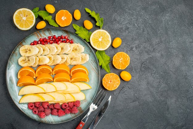 Vista superior de la colección de frutas frescas picadas en una placa azul alrededor de naranjas kumquats y cubiertos en mesa negra