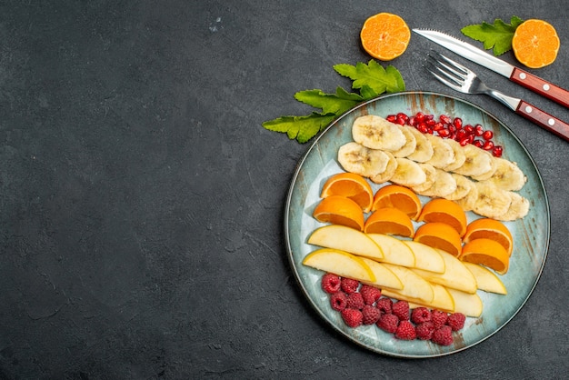 Vista superior de la colección de frutas frescas picadas en un plato azul y cubiertos en mesa negra