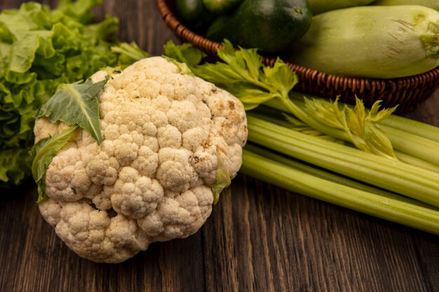 Vista superior de coliflor saludable con verduras como calabacines y pepinos en un balde con apio y lechuga sobre una superficie de madera