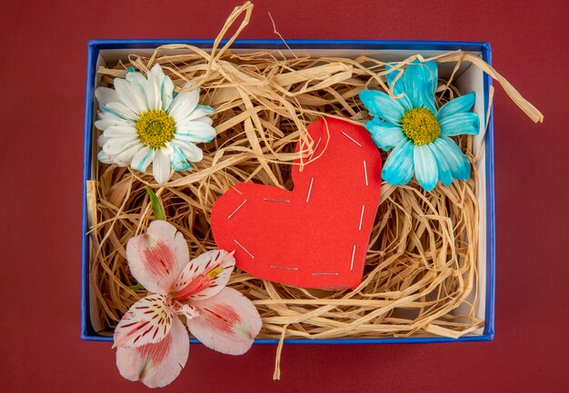 Vista superior de coloridas flores de margarita y alstroemeria rosa con un corazón hecho de papel de color rojo y con paja en una caja de regalo azul sobre mesa roja