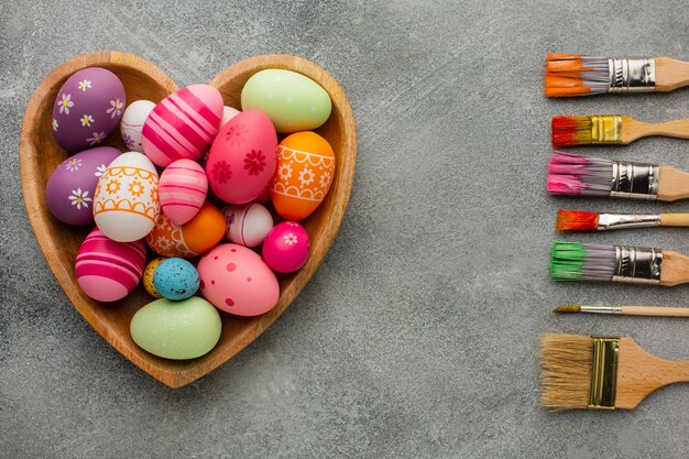 Vista superior de coloridos huevos de pascua en placa en forma de corazón con variedad de pinceles