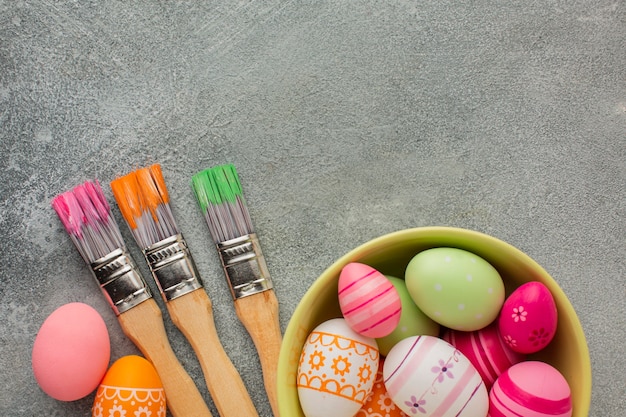 Vista superior de coloridos huevos de pascua en un tazón con pinceles y espacio de copia