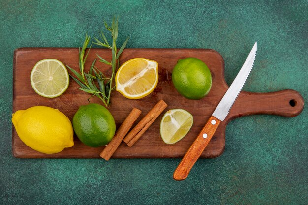 Vista superior de coloridos limones enteros y en rodajas sobre tablero de cocina de madera con ramas de canela con cuchillo en verde