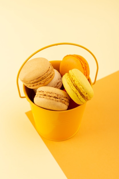 Una vista superior de coloridos macarons franceses deliciosos dentro del cubo amarillo en el escritorio amarillo pastel galleta azúcar dulce