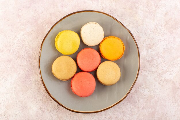 Una vista superior de coloridos macarons franceses dentro de la placa en el escritorio rosa pastel de azúcar galleta dulce