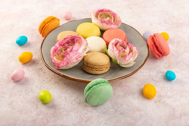 Una vista superior de coloridos macarons franceses dentro de la placa con rosas en el escritorio rosa pastel de azúcar galleta dulce
