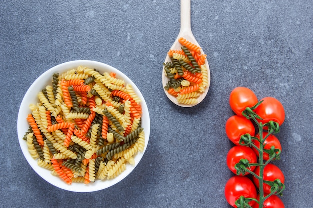 Vista superior coloridos macarrones pasta en un recipiente y una cuchara con tomates en superficie gris. horizontal