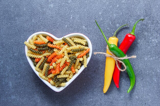 Vista superior coloridos macarrones pasta en un recipiente en forma de corazón con pimienta en la superficie gris. horizontal