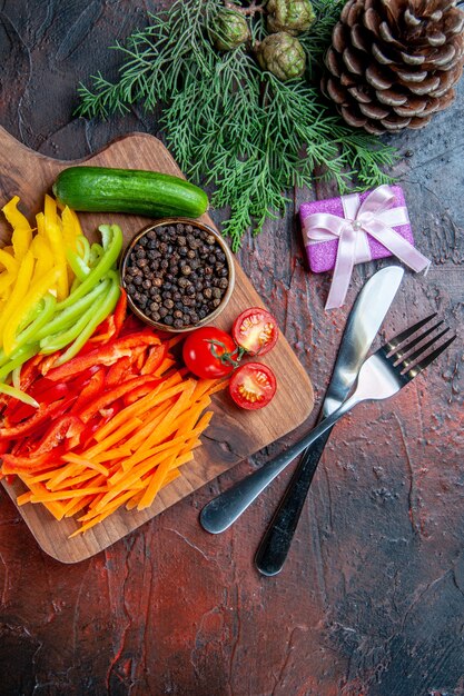 Vista superior de coloridos pimientos cortados, pimienta negra, tomates, pepino, en una tabla de cortar, cuchillo y tenedor, pequeña mesa de color rojo oscuro gifton