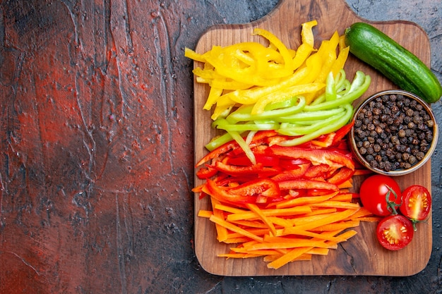 Vista superior de coloridos pimientos cortados, pimienta negra, tomates, pepino en la tabla de cortar en el espacio libre de la mesa rojo oscuro