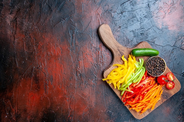 Vista superior de coloridos pimientos cortados, pimienta negra, tomates, pepino en una tabla de cortar en una mesa de color rojo oscuro con espacio de copia