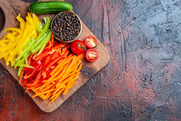 Vista superior de coloridos pimientos cortados, pimienta negra, tomates, pepino en una tabla de cortar en una mesa de color rojo oscuro con espacio de copia