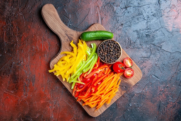 Vista superior de coloridos pimientos cortados, pimienta negra, tomates, pepino en la tabla de cortar en la mesa de color rojo oscuro con espacio de copia