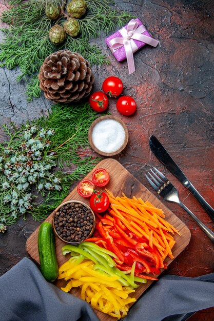 Vista superior de coloridos pimientos cortados, pimienta negra, tomates, pepino en tabla de cortar, regalo pequeño, ramas de pino, cuchillo de sal y tenedor en la mesa de color rojo oscuro