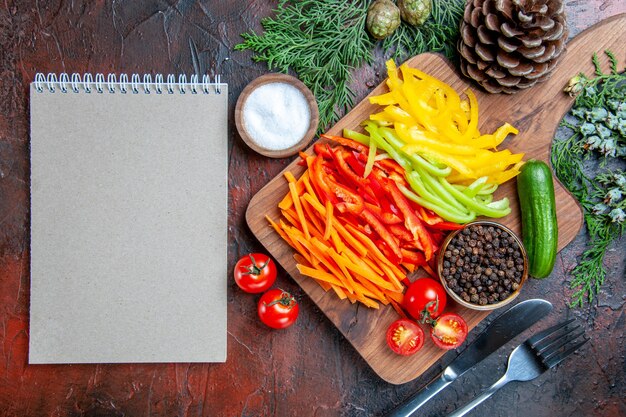 Vista superior de coloridos pimientos cortados, pimienta negra, tomates, pepino, en la tabla de cortar, sal, tenedor y cuchillo, Bloc de notas en la mesa rojo oscuro