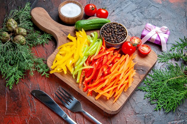 Vista superior de coloridos pimientos cortados, pimienta negra, tomates, pepino en la tabla de cortar, sal, tenedor y cuchillo, pequeño regalo en la mesa de color rojo oscuro