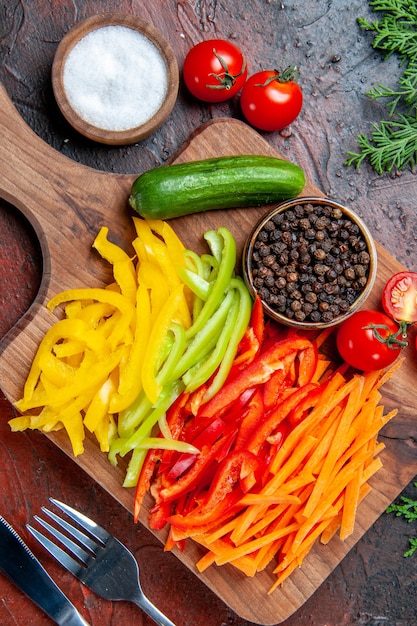 Vista superior de coloridos pimientos cortados, pimienta negra, tomates, pepino en la tabla de cortar, tenedor y cuchillo de sal en la mesa rojo oscuro