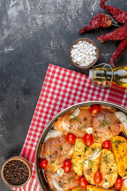 Vista superior de la comida picante de pollo crudo con patatas, verduras en una cacerola sobre una toalla roja pelada y pimientos secos, sal, botella de aceite caída sobre fondo gris