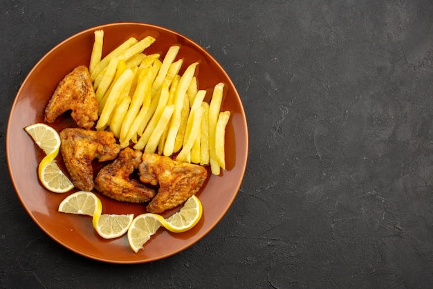 Vista superior de la comida rápida naranja plato de apetitosas papas fritas alitas de pollo y limón en el lado izquierdo de la mesa oscura