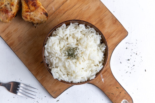 Vista superior de la comida sabrosa de arroz cocido dentro de una olla marrón con aceite y pollo en la superficie blanca