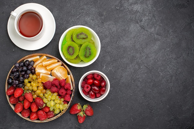 Vista superior de la composición de la fruta fresas uvas frambuesas y mandarinas con una taza de té en el escritorio gris oscuro