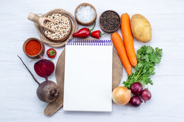 Vista superior de la composición vegetal con verduras frescas, frijoles crudos, zanahorias, bloc de notas y patatas en el escritorio blanco, comida, ensalada de verduras