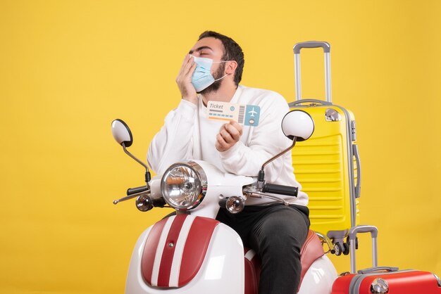 Vista superior del concepto de viaje con un chico de ensueño con máscara médica sentado en una motocicleta con una maleta amarilla y un boleto de explotación