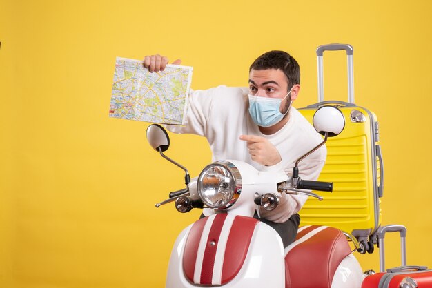 Vista superior del concepto de viaje con un chico joven con máscara médica de pie cerca de la motocicleta con una maleta amarilla y sosteniendo un mapa apuntando hacia arriba